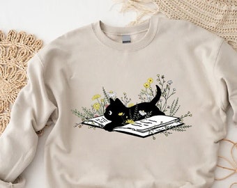 May include: A beige sweatshirt with a black cat reading a book surrounded by flowers. The cat is lying on the open book with its head resting on the pages. The flowers are in shades of yellow, white, and green.