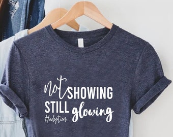 Peut inclure: T-shirt bleu marine avec l'inscription blanche "not showing still glowing" et le hashtag #adoption. Le t-shirt est suspendu à un cintre en bois avec d'autres vêtements, dont une chemise en jean.