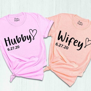 May include: Two pink and peach t-shirts with the words "Hubby" and "Wifey" in black script with a heart and the date "6.27.20" printed on them.