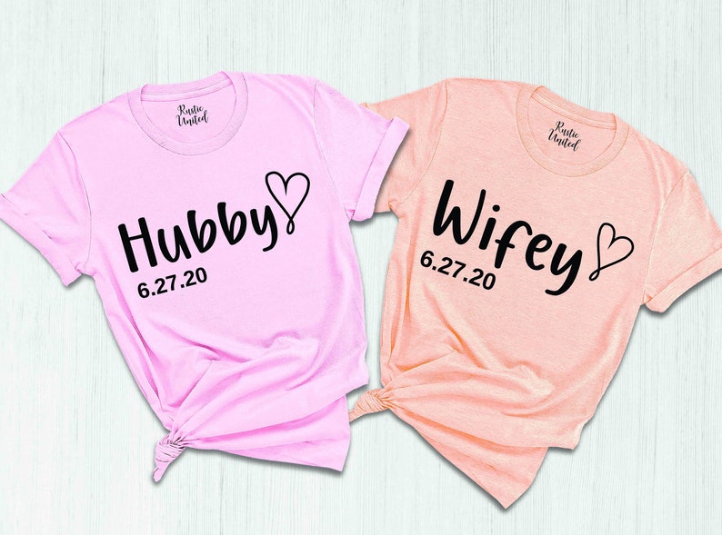 May include: Two pink and peach t-shirts with the words "Hubby" and "Wifey" in black script with a heart and the date "6.27.20" printed on them.