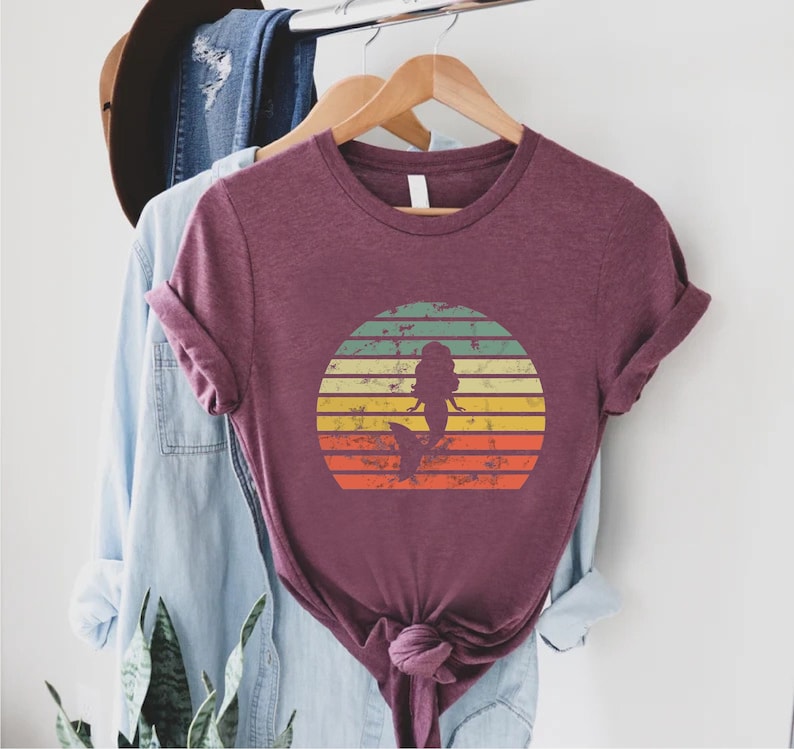 May include: A maroon t-shirt with a vintage-style sunset graphic featuring a mermaid silhouette. The graphic has a distressed look with faded colors and a textured background.