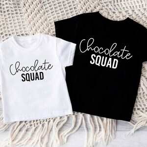 Peut inclure: Deux t-shirts, un blanc et un noir, avec le texte "Chocolate Squad" imprim&eacute; en blanc sur le devant.