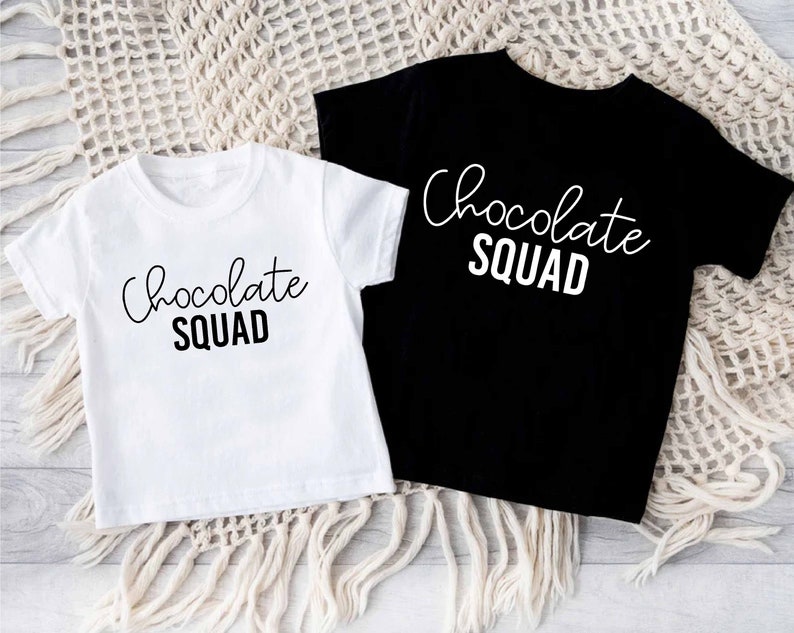 Peut inclure: Deux t-shirts, un blanc et un noir, avec le texte "Chocolate Squad" imprim&eacute; en blanc sur le devant.
