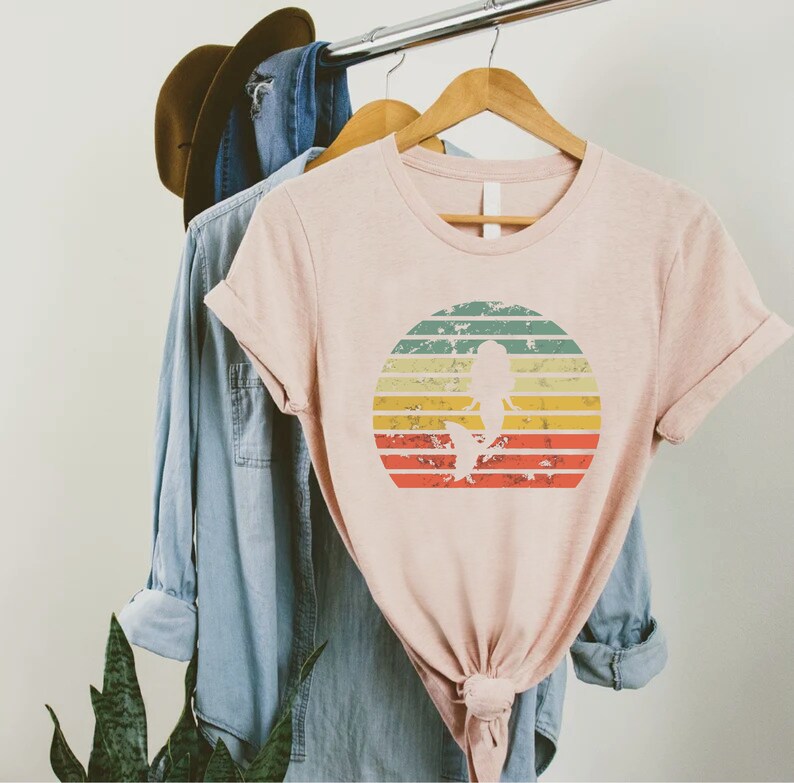May include: A pink t-shirt with a vintage-style graphic of a mermaid silhouette in front of a sunset. The graphic has a distressed look with faded colors and a white outline.