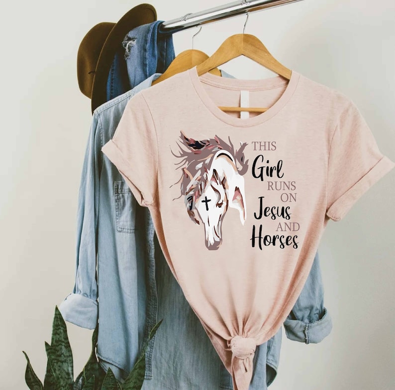 May include: A pink t-shirt with a graphic of a horse's head with a cross on its forehead. The text "This Girl Runs On Jesus And Horses" is printed on the shirt.