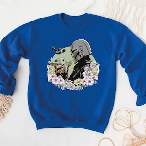 May include: A royal blue sweatshirt featuring a graphic print of a Mandalorian and Grogu, surrounded by flowers. The Mandalorian is wearing his helmet and holding Grogu in his arms.