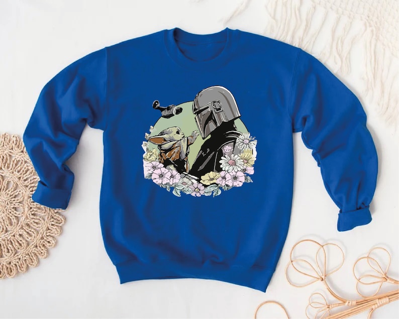 May include: A royal blue sweatshirt featuring a graphic print of a Mandalorian and Grogu, surrounded by flowers. The Mandalorian is wearing his helmet and holding Grogu in his arms.