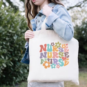 Puede incluir: Una bolsa de lona blanca con un dise&ntilde;o retro colorido que dice "NURSE" con flores y un coraz&oacute;n.