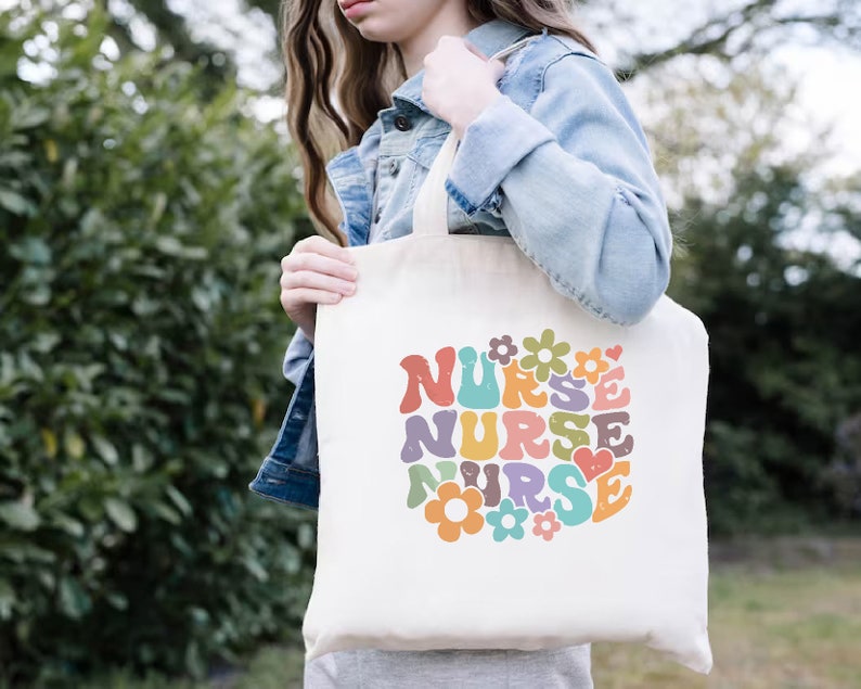 Puede incluir: Una bolsa de lona blanca con un dise&ntilde;o retro colorido que dice "NURSE" con flores y un coraz&oacute;n.