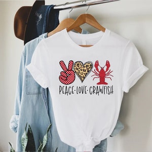 May include: White t-shirt with a peace sign, heart, and crawfish graphic. The peace sign is red with white polka dots, the heart is leopard print, and the crawfish is red. The text "Peace Love Crawfish" is printed below the graphic.