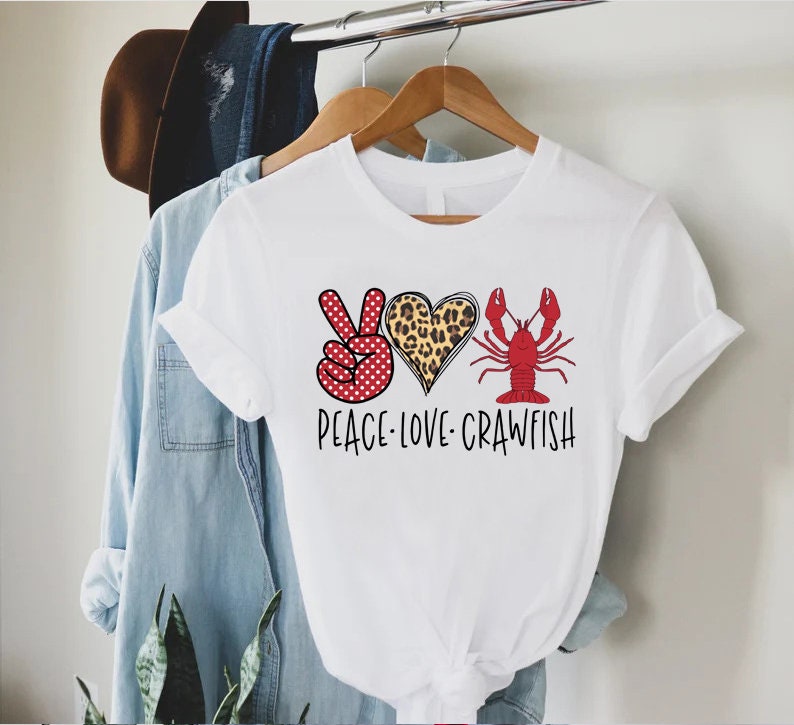 May include: White t-shirt with a peace sign, heart, and crawfish graphic. The peace sign is red with white polka dots, the heart is leopard print, and the crawfish is red. The text "Peace Love Crawfish" is printed below the graphic.