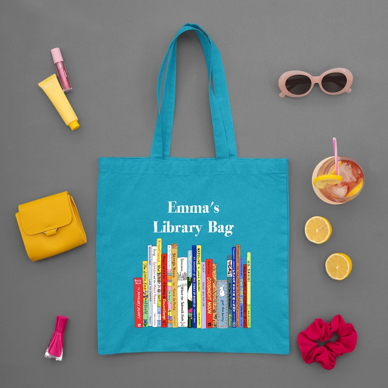 May include: A turquoise tote bag with the text "Emma's Library Bag" and a graphic of colorful books. The bag is surrounded by a yellow wallet, sunglasses, a drink, and other accessories on a gray surface.