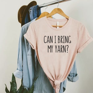 May include: A light pink t-shirt with the text "CAN I BRING MY YARN?" in black lettering. The shirt is hanging on a wooden hanger with a denim shirt and a brown hat in the background. The shirt is tied at the bottom.