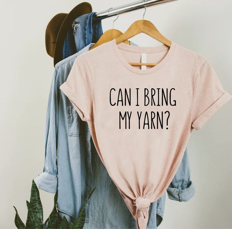 May include: A light pink t-shirt with the text "CAN I BRING MY YARN?" in black lettering. The shirt is hanging on a wooden hanger with a denim shirt and a brown hat in the background. The shirt is tied at the bottom.
