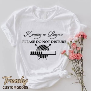 May include: White t-shirt with black text that reads "Knitting in Progress, Please Do Not Disturb". A black and white illustration of yarn and knitting needles is below the text with a progress bar underneath.
