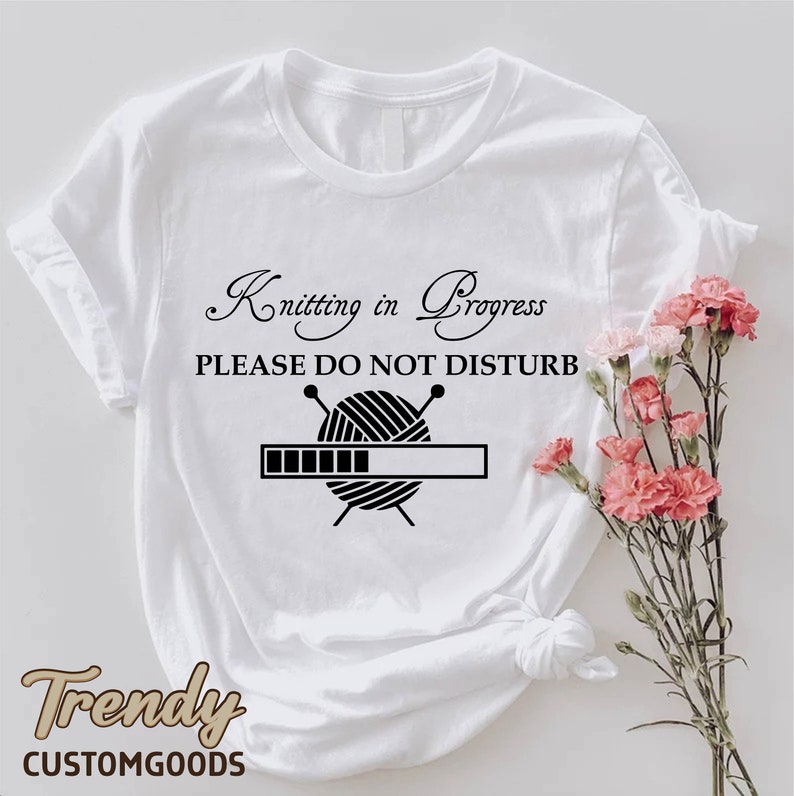 May include: White t-shirt with black text that reads "Knitting in Progress, Please Do Not Disturb". A black and white illustration of yarn and knitting needles is below the text with a progress bar underneath.
