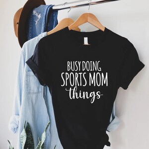 May include: Black t-shirt with white text that reads "Busy doing sports mom things".