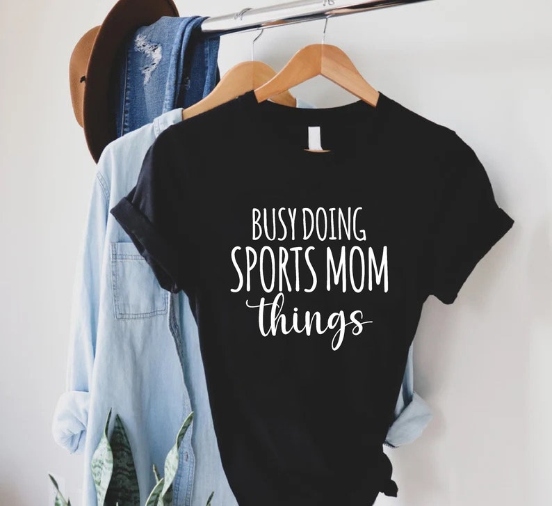 May include: Black t-shirt with white text that reads "Busy doing sports mom things".