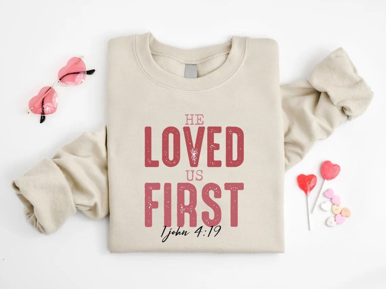 Puede incluir: Sudadera beige con el texto "HE LOVED US FIRST 1 John 4:19" en letras rojas. La sudadera est&aacute; doblada y colocada sobre una superficie blanca con gafas de sol de coraz&oacute;n rosa y corazones de caramelo.