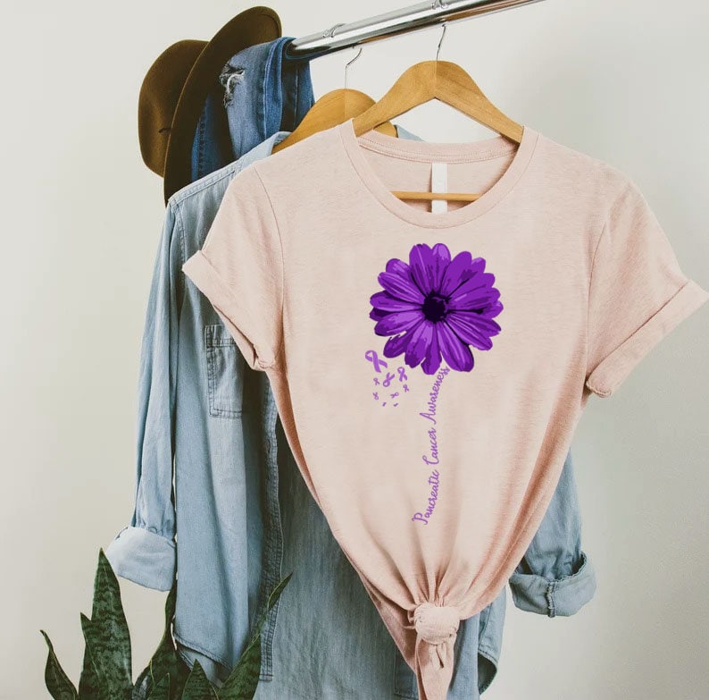 K&ouml;nnte beinhalten: Ein rosa T-Shirt mit einer lila Blume und lila B&auml;ndern. Der Text "Pancreatic Cancer Awareness" ist vertikal auf der rechten Seite des Shirts geschrieben.