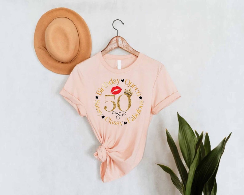 May include: Pink t-shirt with a gold glitter design that says "Birthday Queen 50 Sassy Classy Fabulous" with a red lip and crown graphic.