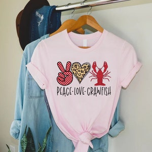 May include: Pink t-shirt with a peace sign, heart, and crawfish graphic. The peace sign is red with white polka dots, the heart is leopard print, and the crawfish is red. The text "Peace Love Crawfish" is printed below the graphic.