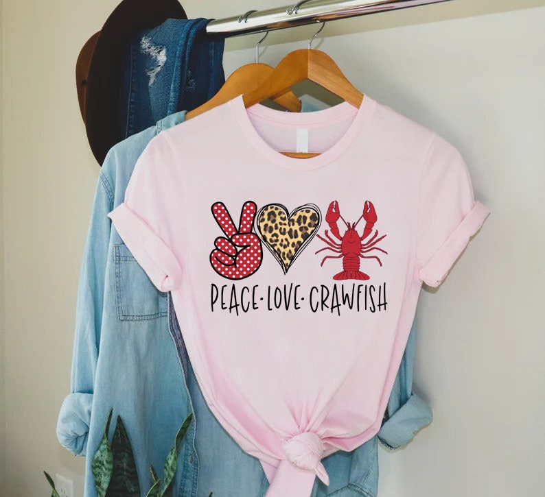 May include: Pink t-shirt with a peace sign, heart, and crawfish graphic. The peace sign is red with white polka dots, the heart is leopard print, and the crawfish is red. The text "Peace Love Crawfish" is printed below the graphic.