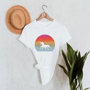 K&ouml;nnte beinhalten: Wei&szlig;es T-Shirt mit einem Retro-Design, das eine Einhorn-Silhouette in einem farbenfrohen Sonnenuntergang zeigt. Der Sonnenuntergang hat horizontale Streifen in Rosa, Orange, Gelb und Blau. Ein brauner Hut h&auml;ngt an der Wand hinter dem Shirt.