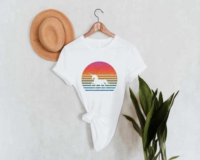 K&ouml;nnte beinhalten: Wei&szlig;es T-Shirt mit einem Retro-Design, das eine Einhorn-Silhouette in einem farbenfrohen Sonnenuntergang zeigt. Der Sonnenuntergang hat horizontale Streifen in Rosa, Orange, Gelb und Blau. Ein brauner Hut h&auml;ngt an der Wand hinter dem Shirt.