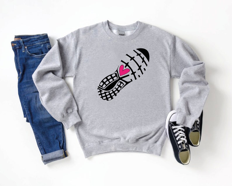 Peut inclure: Sweat-shirt gris avec un imprim&eacute; de chaussure de course noire et blanche avec un c&oelig;ur rose. Le sweat-shirt est pos&eacute; sur une surface blanche avec un jean bleu et des baskets noires.