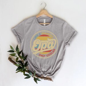 May include: Grey t-shirt featuring a retro-style graphic that reads "Like a Grandpa, Only Cooler" with the word "Opa" in a large, stylised font.
