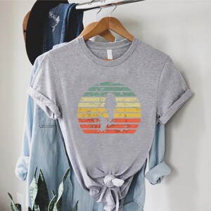 May include: A gray t-shirt with a vintage-style sunset graphic featuring a silhouette of a person. The sunset is in shades of orange, yellow, green, and blue.