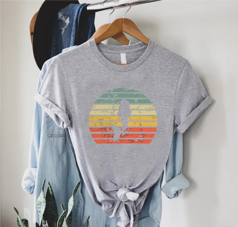 May include: A gray t-shirt with a vintage-style sunset graphic featuring a silhouette of a person. The sunset is in shades of orange, yellow, green, and blue.