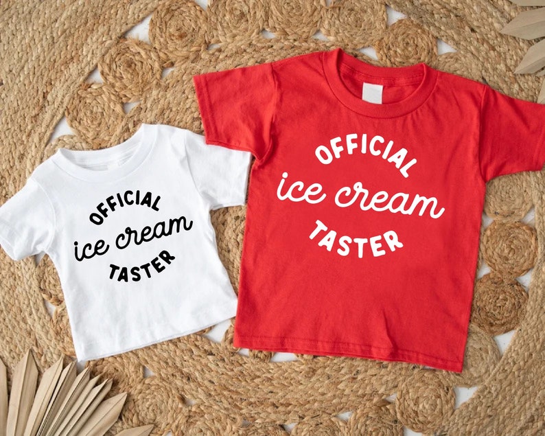 Puede incluir: Dos camisetas blancas con texto negro que dice "OFFICIAL ice cream TASTER" sobre un fondo tejido marr&oacute;n.