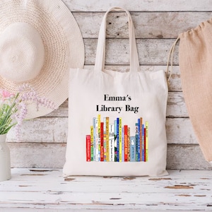 May include: A cream-colored tote bag with the text "Emma's Library Bag" above a colorful stack of books. The bag has long handles and is made of a sturdy fabric. A white sun hat and a vase of flowers are in the background.