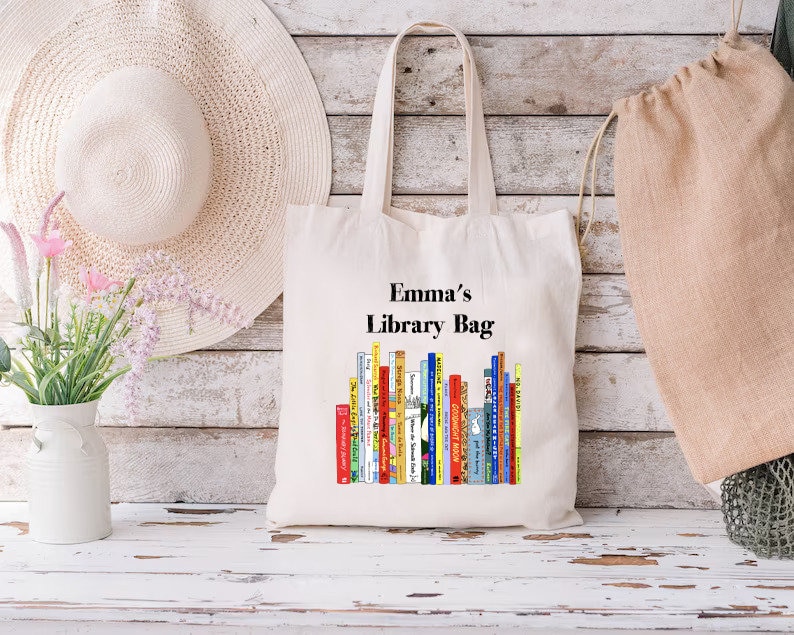 May include: A cream-colored tote bag with the text "Emma's Library Bag" above a colorful stack of books. The bag has long handles and is made of a sturdy fabric. A white sun hat and a vase of flowers are in the background.
