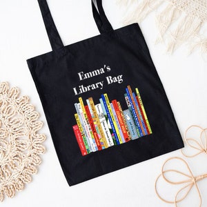 May include: Black tote bag with the text "Emma's Library Bag" above a colorful illustration of a stack of books. The bag has two black handles and is made of a sturdy fabric.