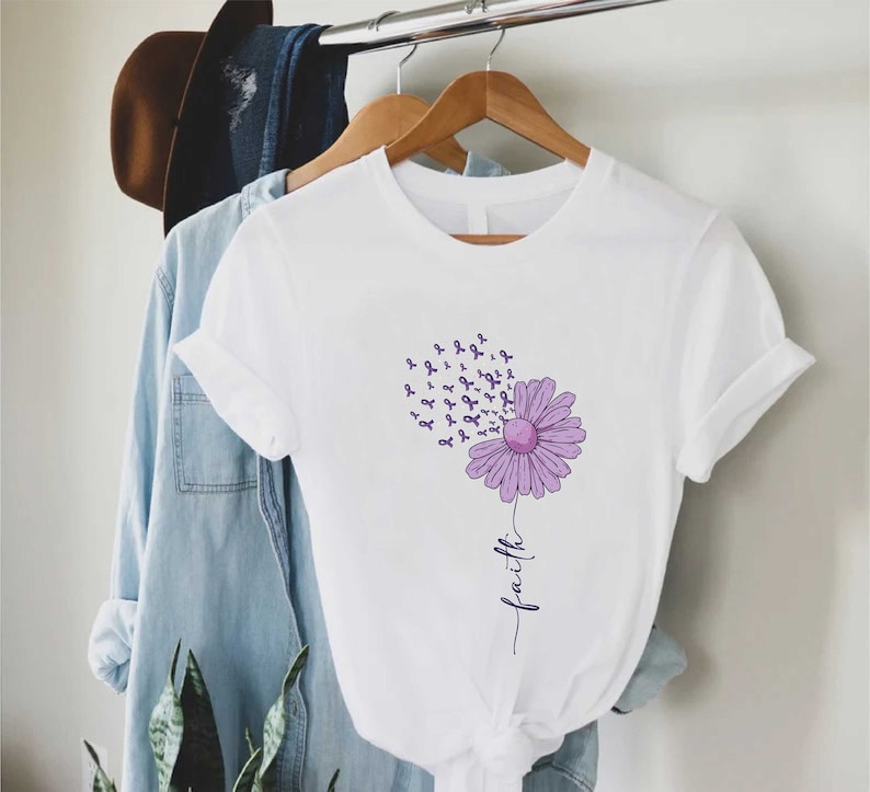May include: White t-shirt with a purple flower design. The flower has purple ribbons coming out of it. The word "faith" is written vertically below the flower.