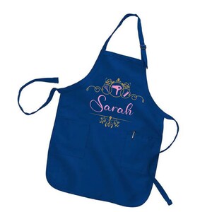Puede incluir: Un delantal azul marino con dos bolsillos delanteros y un bolsillo para bol&iacute;grafo. El delantal presenta el nombre "Sarah" en escritura rosa, junto con herramientas de peluquer&iacute;a doradas y adornos decorativos. El delantal tiene una correa ajustable para el cuello.