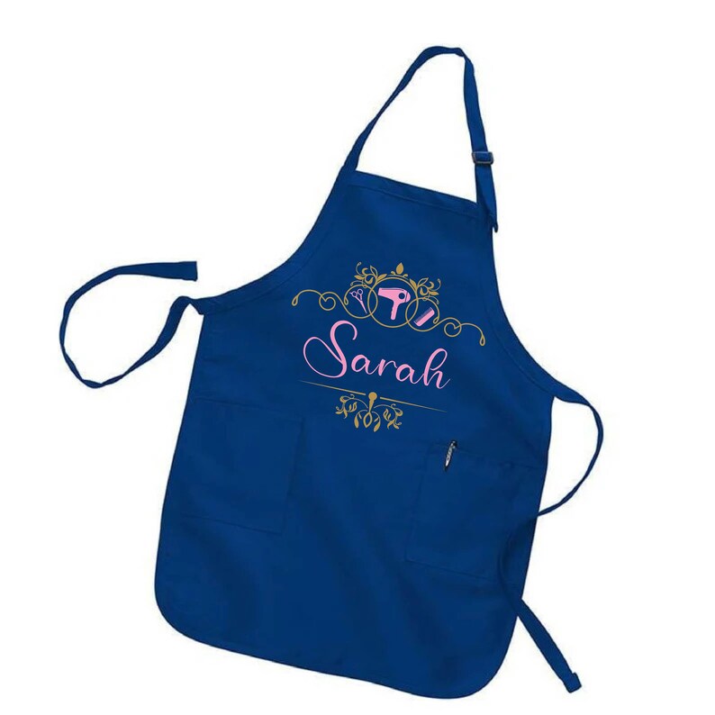 Puede incluir: Un delantal azul marino con dos bolsillos delanteros y un bolsillo para bol&iacute;grafo. El delantal presenta el nombre "Sarah" en escritura rosa, junto con herramientas de peluquer&iacute;a doradas y adornos decorativos. El delantal tiene una correa ajustable para el cuello.
