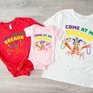 Może przedstawiać: Trzy koszulki w kolorze czerwonym, r&oacute;żowym i białym z napisem "Come at me Breaux" i kresk&oacute;wkowym rakiem w kapeluszu błazna i koralikami Mardi Gras.