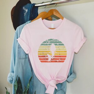 May include: Pink t-shirt with a vintage-style graphic of a mermaid silhouette in a sunset. The graphic has a distressed look with faded colors and a white outline.