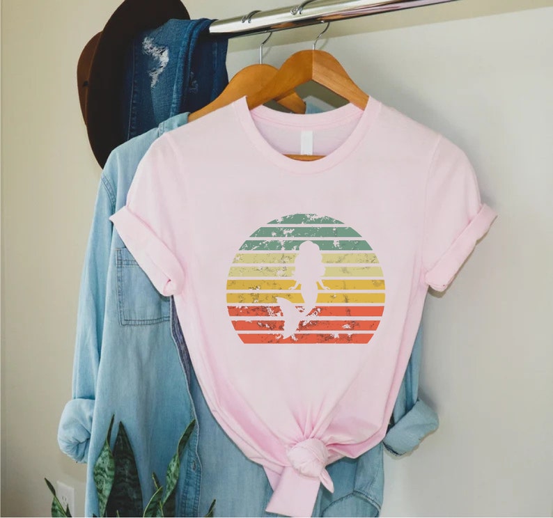 May include: Pink t-shirt with a vintage-style graphic of a mermaid silhouette in a sunset. The graphic has a distressed look with faded colors and a white outline.