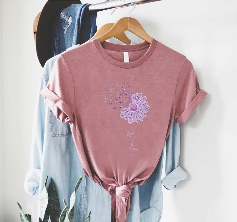 May include: A mauve t-shirt with a purple flower graphic and the word "faith" written vertically below the flower. The flower is made up of small purple and gold shapes.