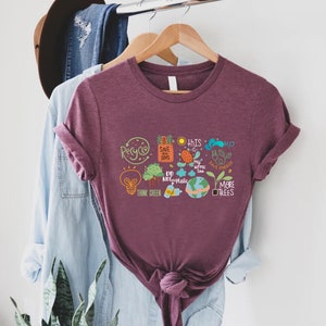 Puede incluir: Una camiseta color granate con un estampado gr&aacute;fico blanco de una bombilla, &aacute;rboles, un globo terr&aacute;queo y otros iconos ambientales. El texto de la camiseta dice "Recycle", "Save our Seas", "This Home Too", "Do Not Use Plastic", "Think Green", "Save Energy" y "More Trees".