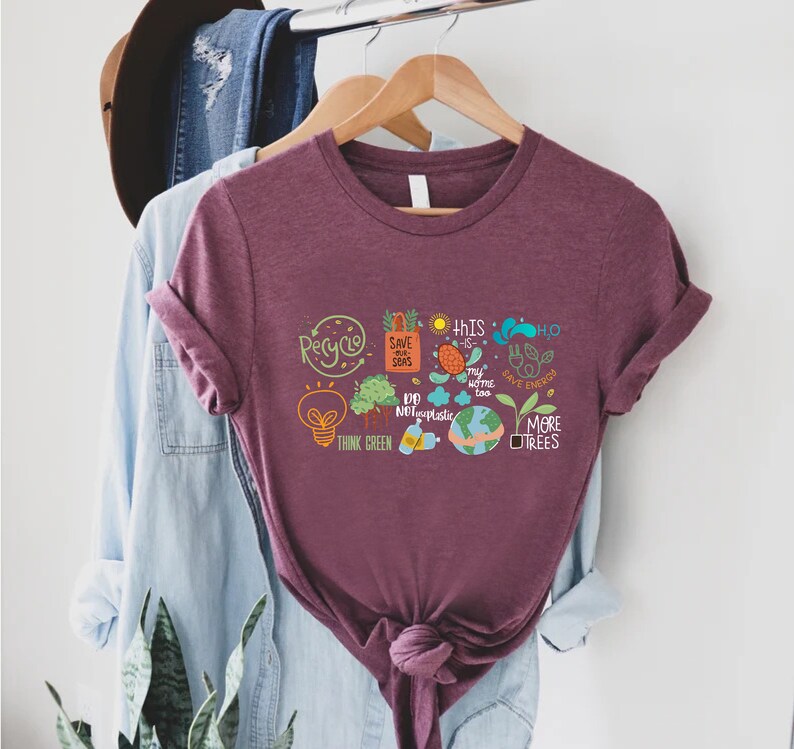 Puede incluir: Una camiseta color granate con un estampado gr&aacute;fico blanco de una bombilla, &aacute;rboles, un globo terr&aacute;queo y otros iconos ambientales. El texto de la camiseta dice "Recycle", "Save our Seas", "This Home Too", "Do Not Use Plastic", "Think Green", "Save Energy" y "More Trees".