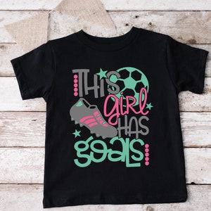 May include: Black t-shirt with a green and pink graphic that says "This girl has goals!" with a soccer ball and soccer cleats.