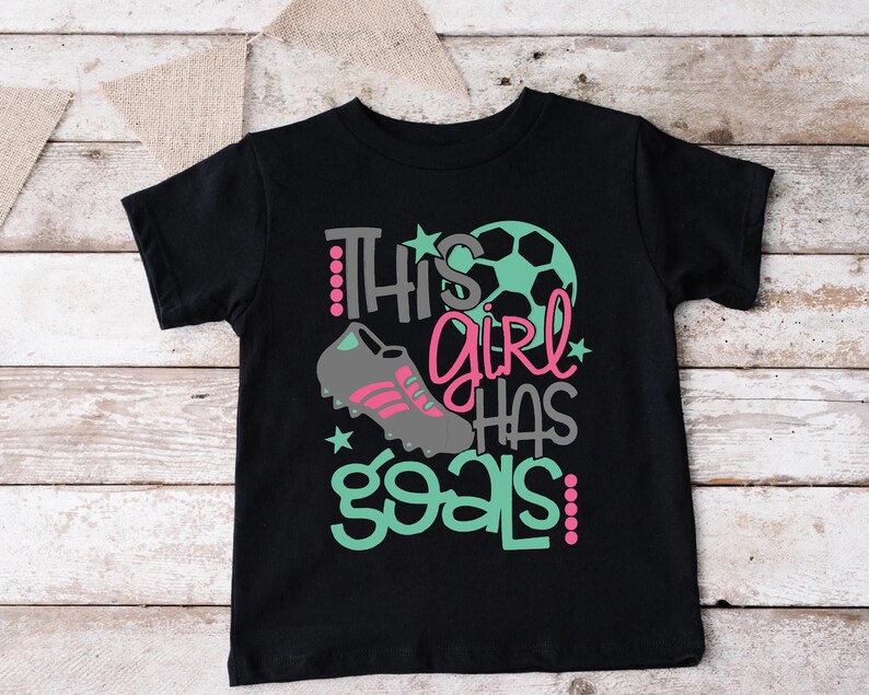 May include: Black t-shirt with a green and pink graphic that says "This girl has goals!" with a soccer ball and soccer cleats.