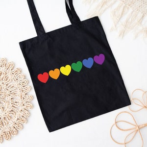 May include: Black tote bag with a rainbow of hearts printed on the front. The hearts are in the order of the rainbow, red, orange, yellow, green, blue, and purple.