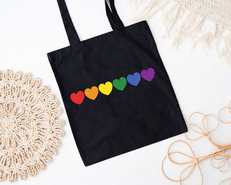 May include: Black tote bag with a rainbow of hearts printed on the front. The hearts are in the order of the rainbow, red, orange, yellow, green, blue, and purple.